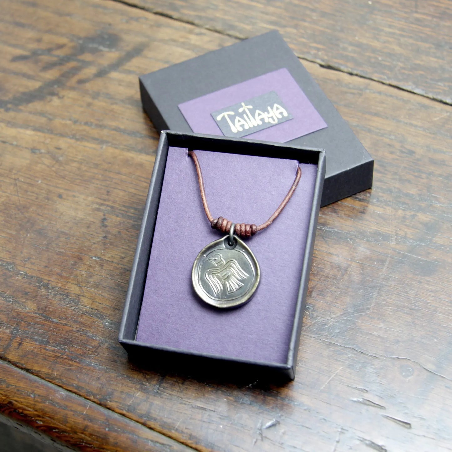 Image of the black, purple and gold branded card jewellery box holding a brass brushed Viking Raven Iron Coin Necklace. Norse Raven medallion pendant that can be personalised with silver inlay or a personal engraving.