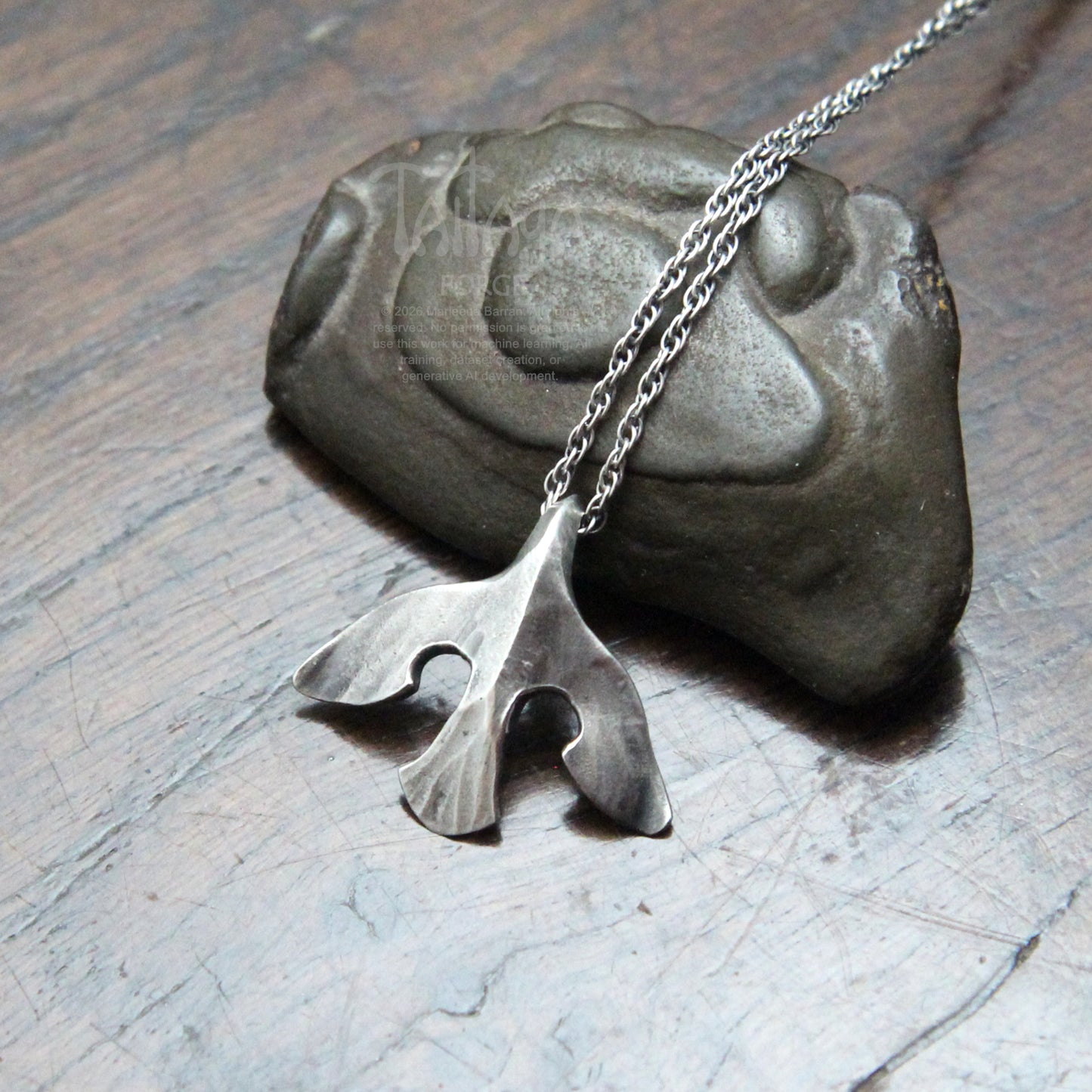 A silver raven pendant necklace with a stylized design, draped over a dark stone on a scratched gray surface.
