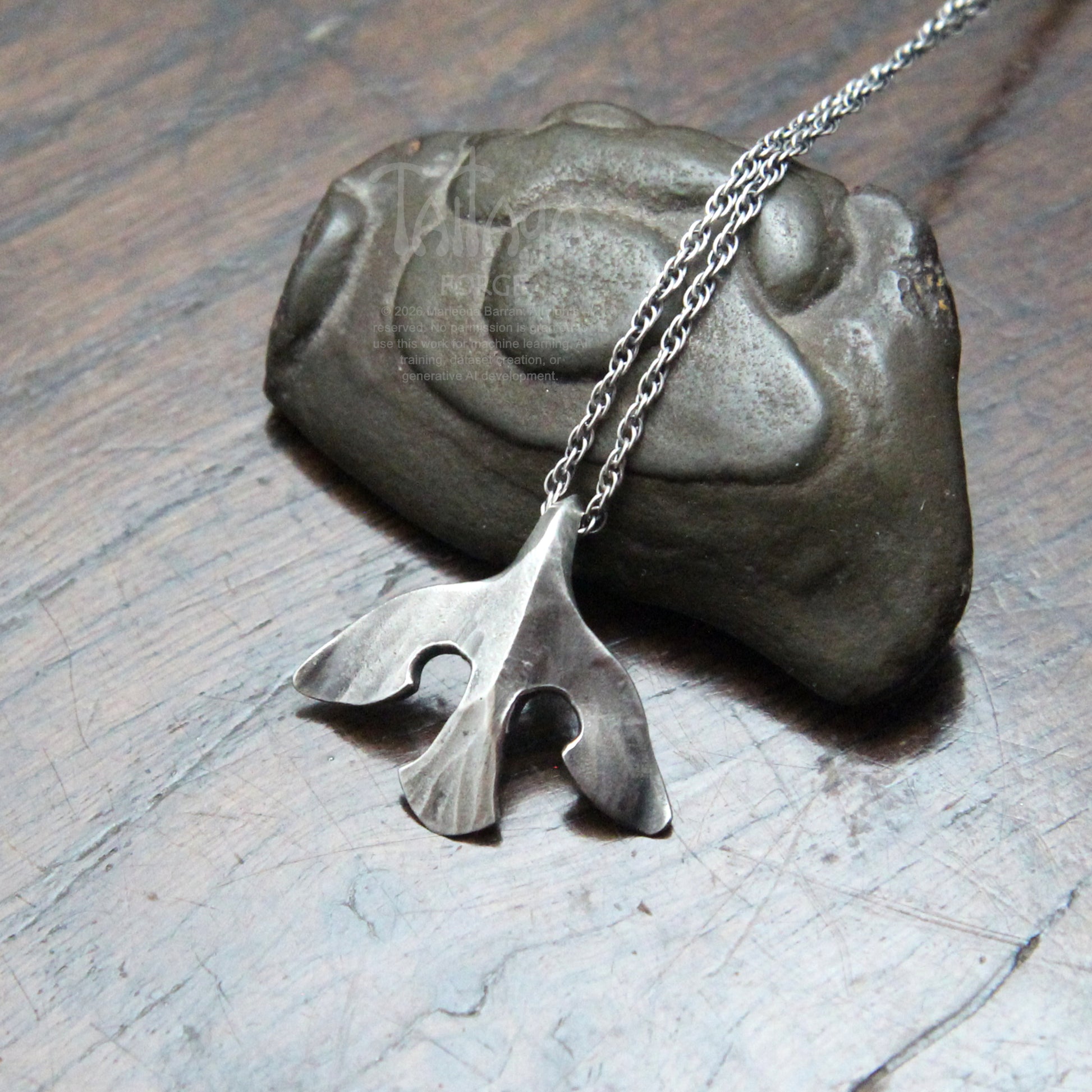 A silver raven pendant necklace with a stylized design, draped over a dark stone on a scratched gray surface.