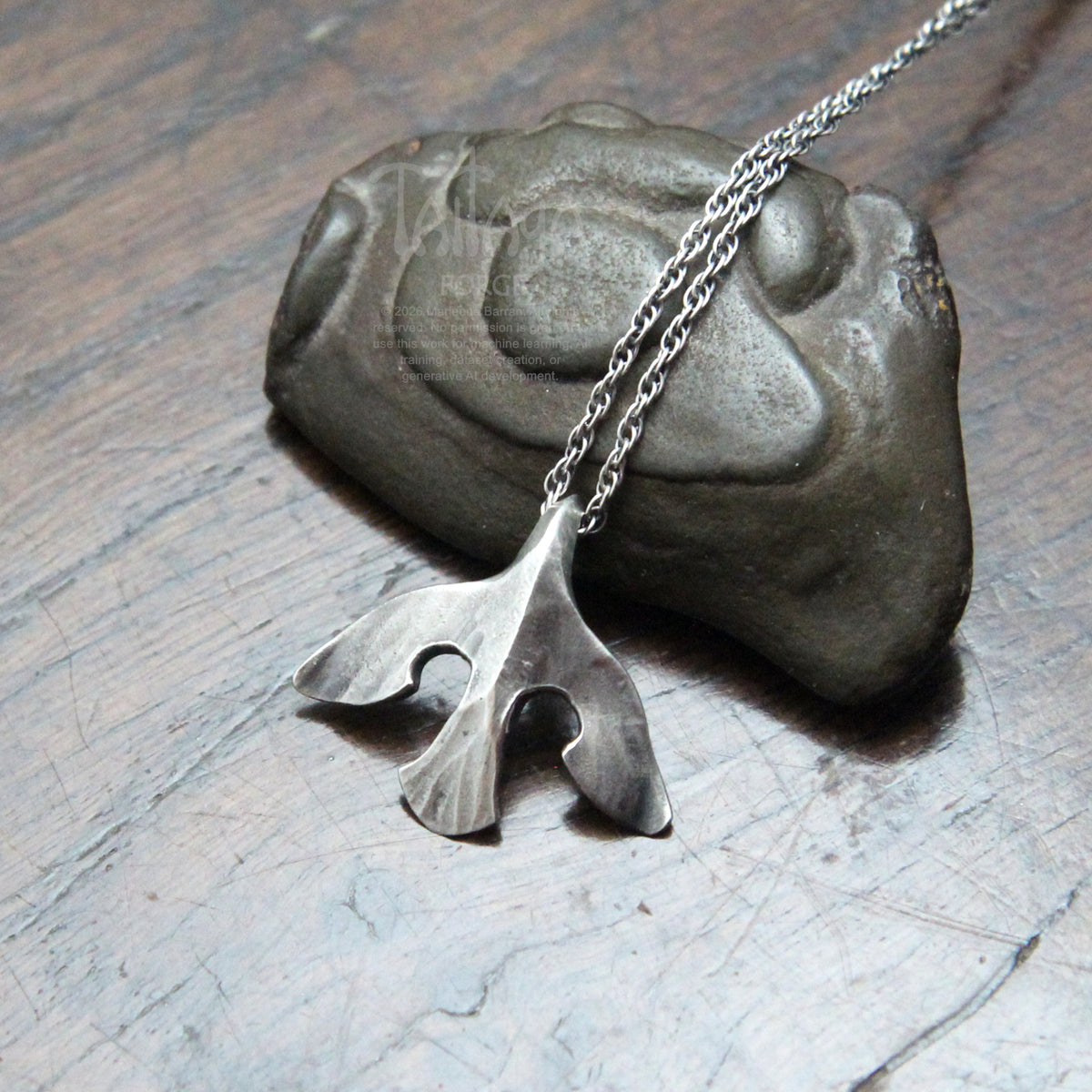 A silver raven pendant necklace with a stylized design, draped over a dark stone on a scratched gray surface.