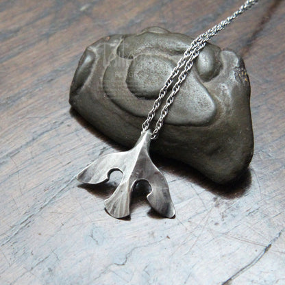 A silver raven pendant necklace with a stylized design, draped over a dark stone on a scratched gray surface.