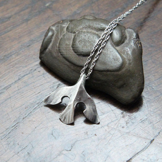 A silver raven pendant necklace with a stylized design, draped over a dark stone on a scratched gray surface.
