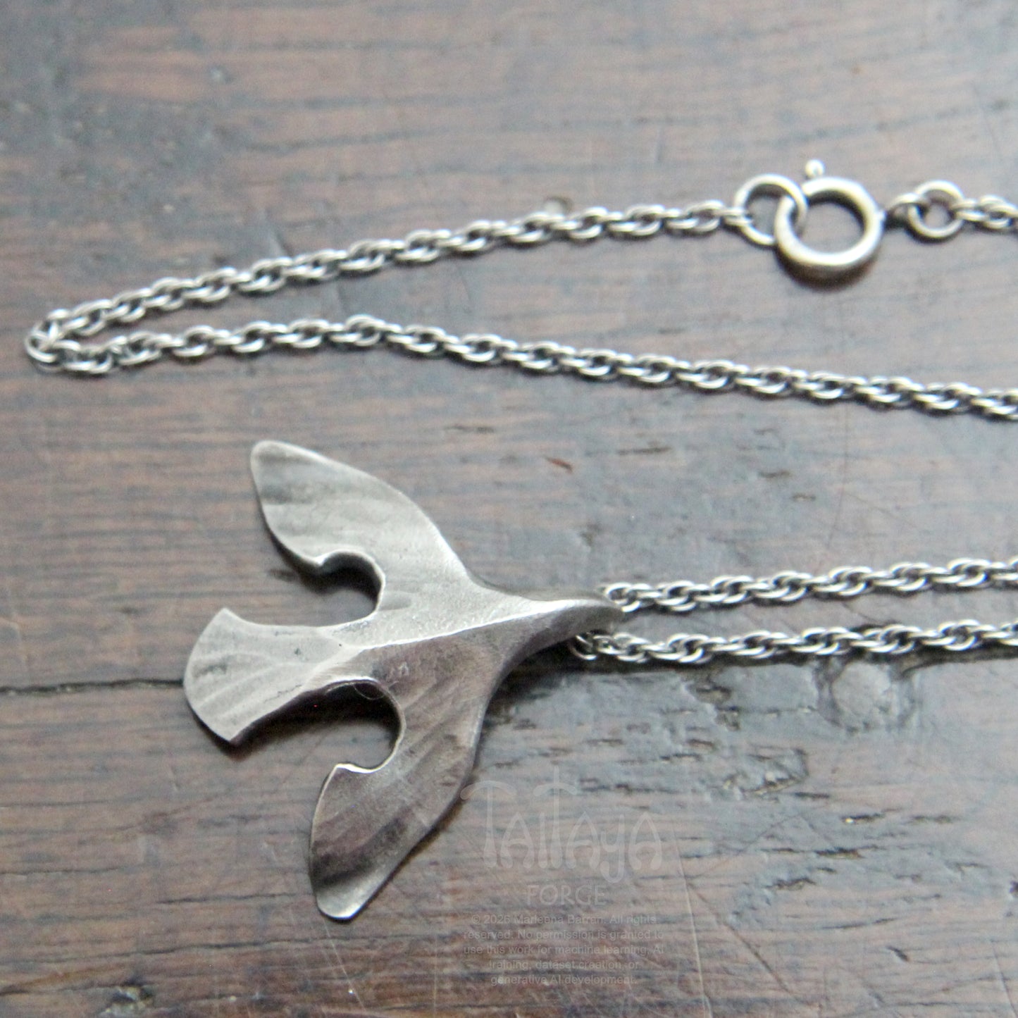 A silver raven pendant necklace featuring a stylized bird on a delicate chain, displayed on a dark wooden surface. Showing Spring ring clasp. 