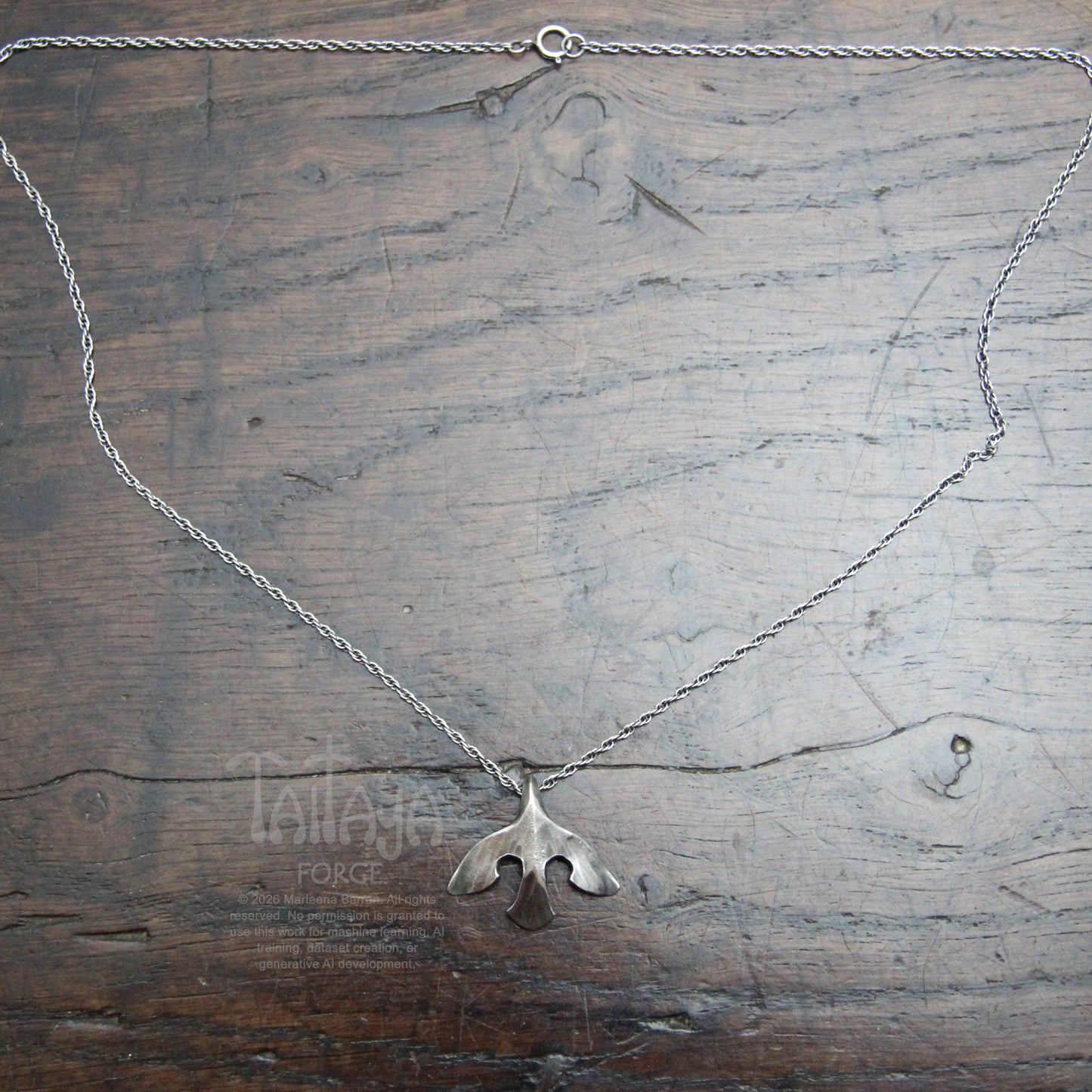 A silver necklace with a smallstylised raven shaped pendant, laid flat on a rustic dark wood background. wood.