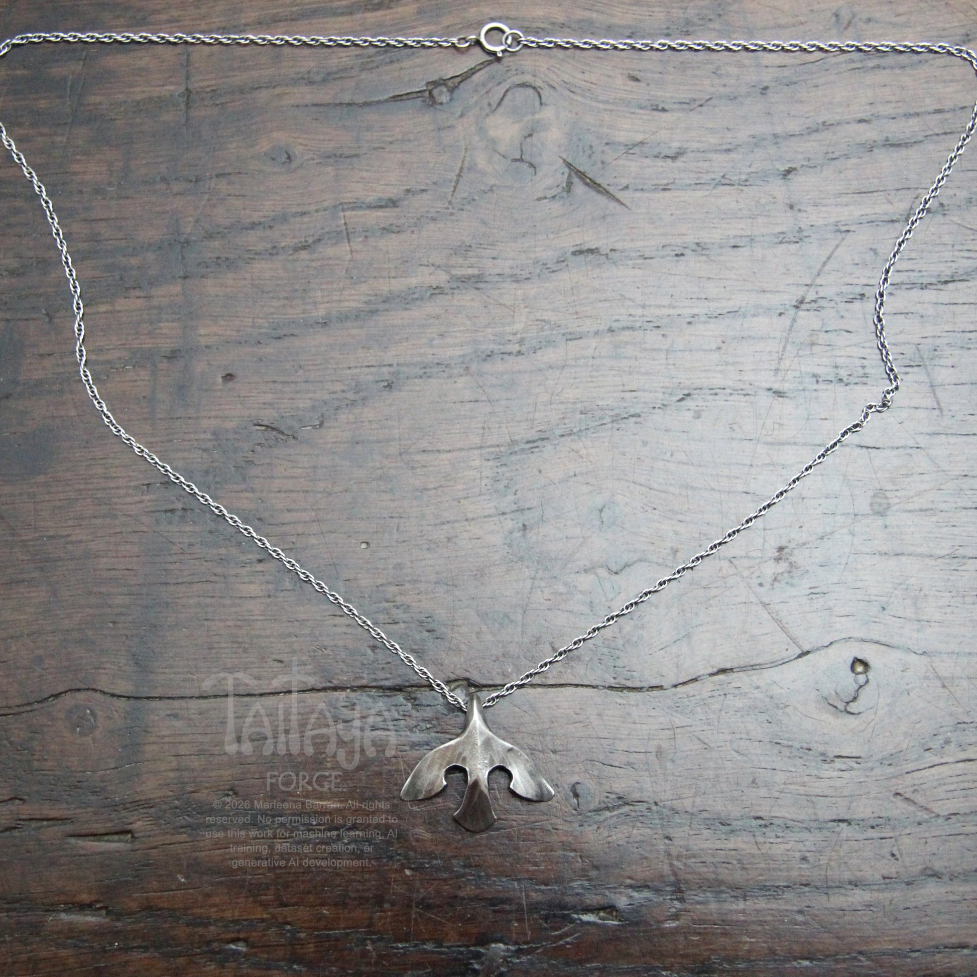 A silver necklace with a smallstylised raven shaped pendant, laid flat on a rustic dark wood background. wood.