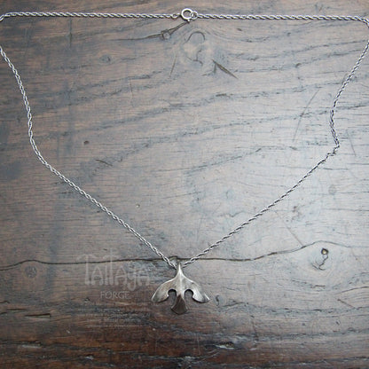 A silver necklace with a smallstylised raven shaped pendant, laid flat on a rustic dark wood background. wood.