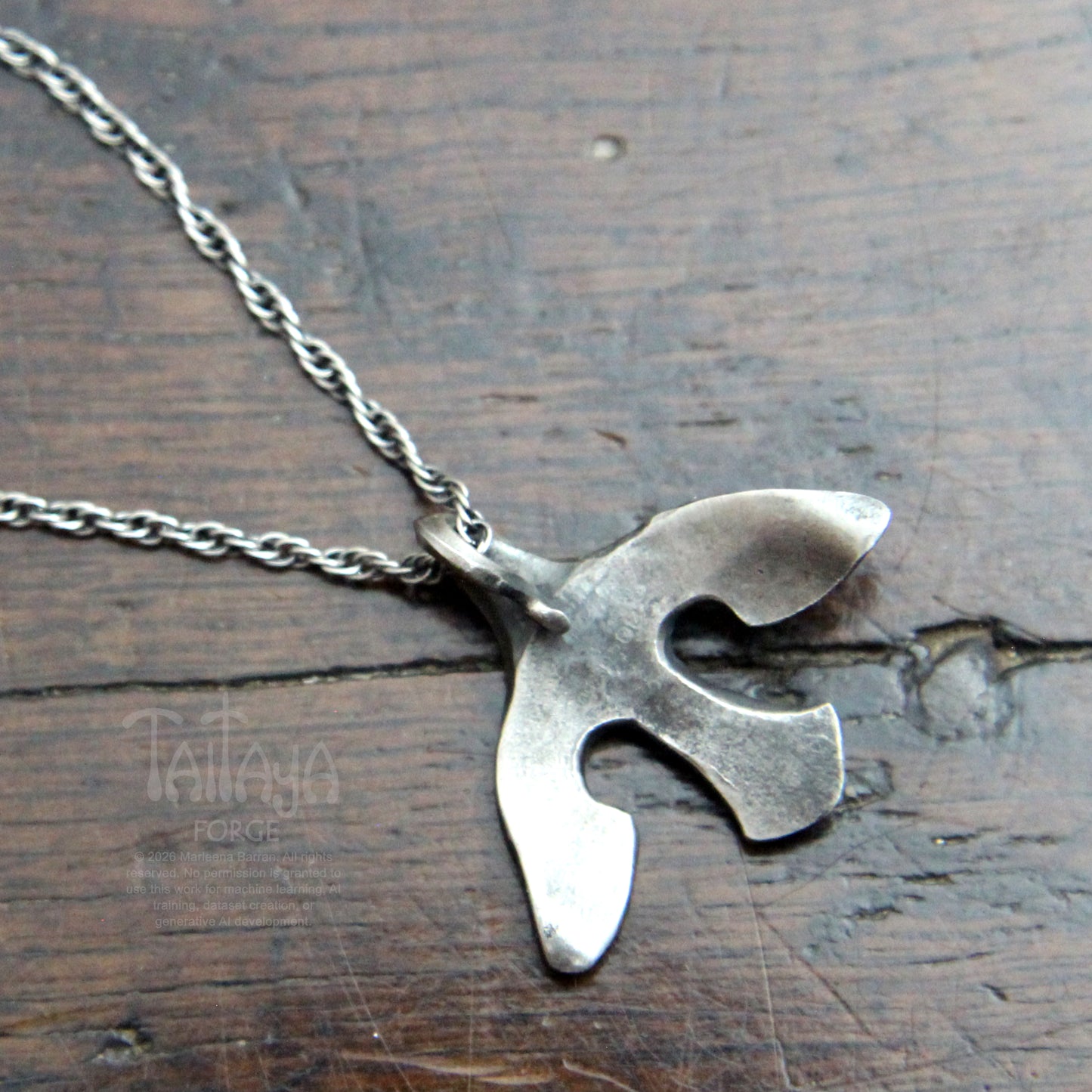 The back of a silver pendant necklace with a stylized, organic bird-like silhouette, resting  on a dark brown wooden surface. The rope chain is partially visible in the upper left corner.