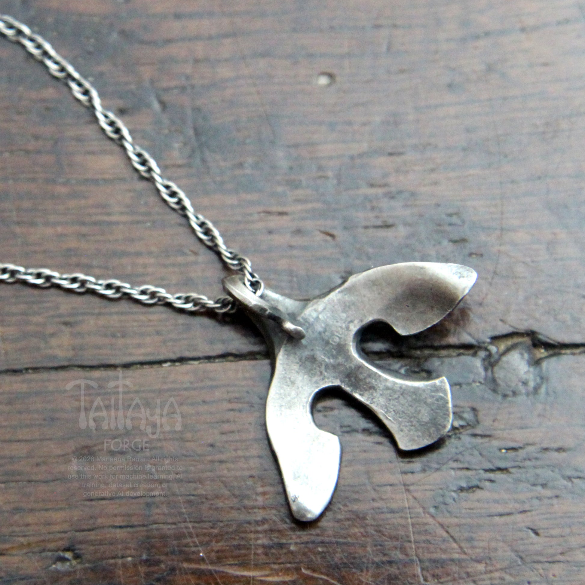 The back of a silver pendant necklace with a stylized, organic bird-like silhouette, resting  on a dark brown wooden surface. The rope chain is partially visible in the upper left corner.