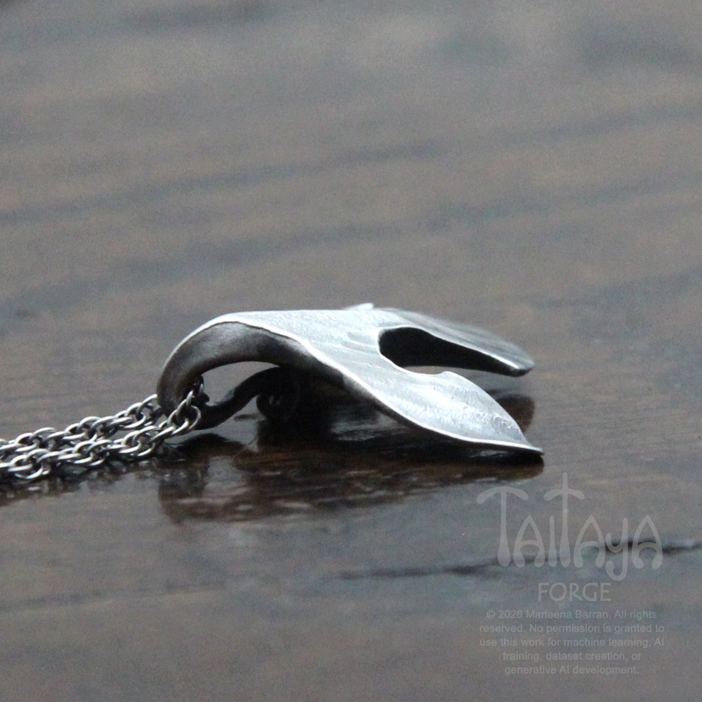 A side photo of a silver raven pendant necklace with a stylized, organic curved shape beak resting on a dark, reflective wooden surface. The chain is visible on the left.