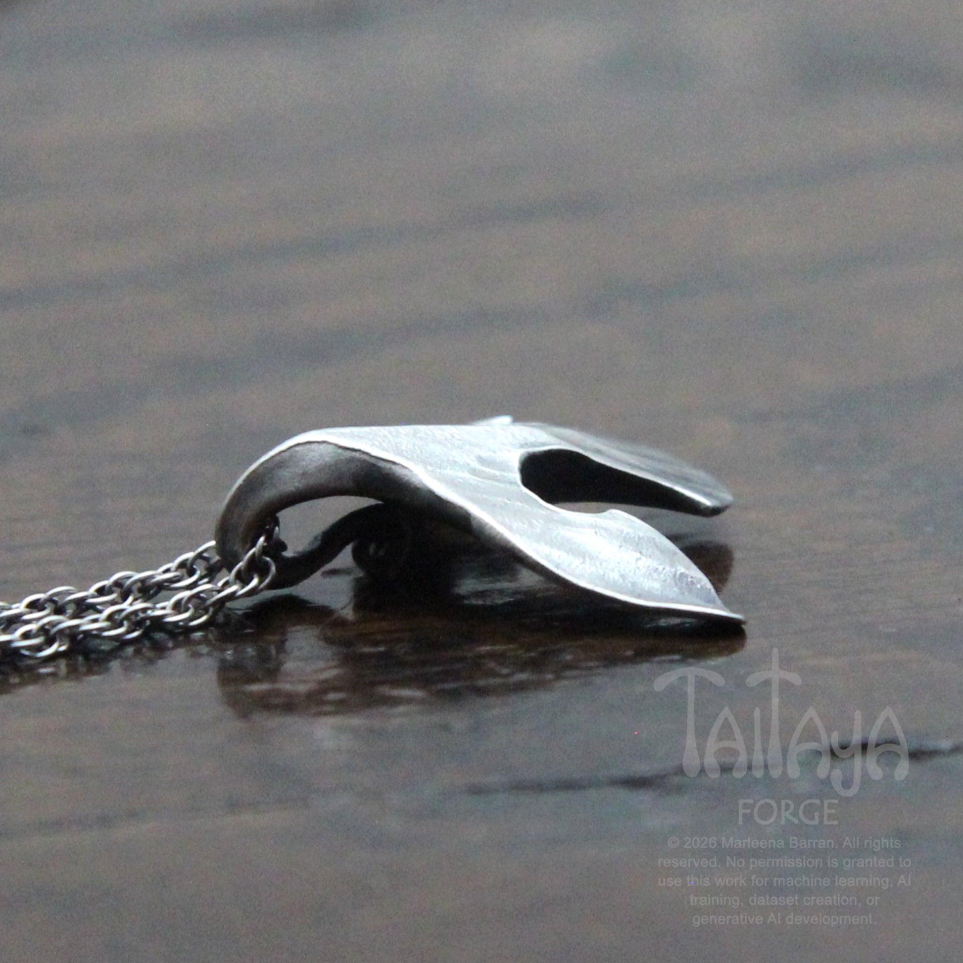 A side photo of a silver raven pendant necklace with a stylized, organic curved shape beak resting on a dark, reflective wooden surface. The chain is visible on the left.