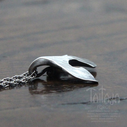 A side photo of a silver raven pendant necklace with a stylized, organic curved shape beak resting on a dark, reflective wooden surface. The chain is visible on the left.