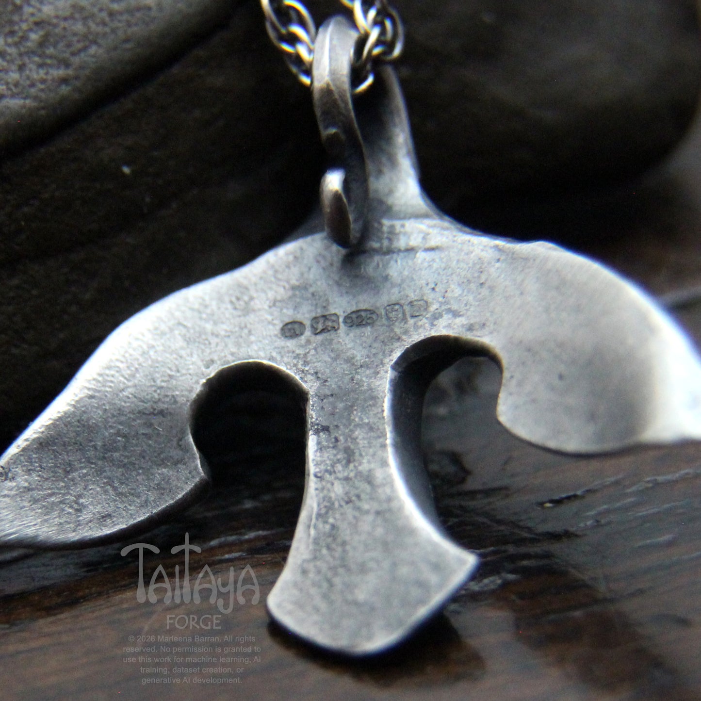 A close-up of a the back of stylized silver raven pendant with UK hallmarks . The pendant is displayed on a dark wooden surface with a dark stone in the background.