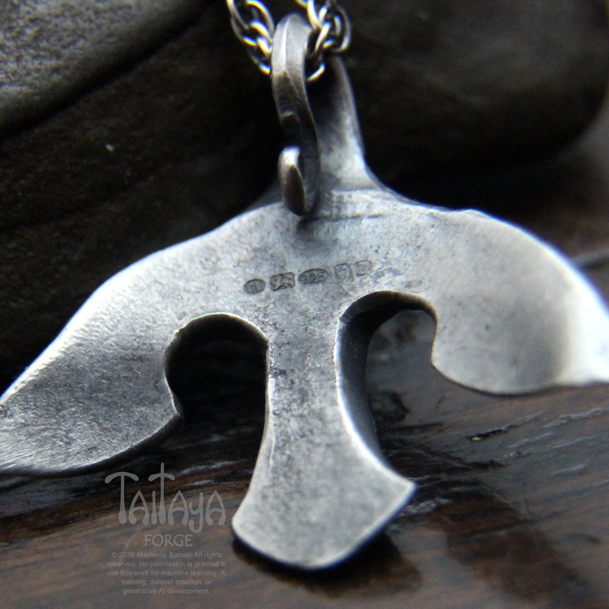 A close-up of a the back of stylized silver raven pendant with UK hallmarks . The pendant is displayed on a dark wooden surface with a dark stone in the background.