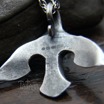 A close-up of a the back of stylized silver raven pendant with UK hallmarks . The pendant is displayed on a dark wooden surface with a dark stone in the background.