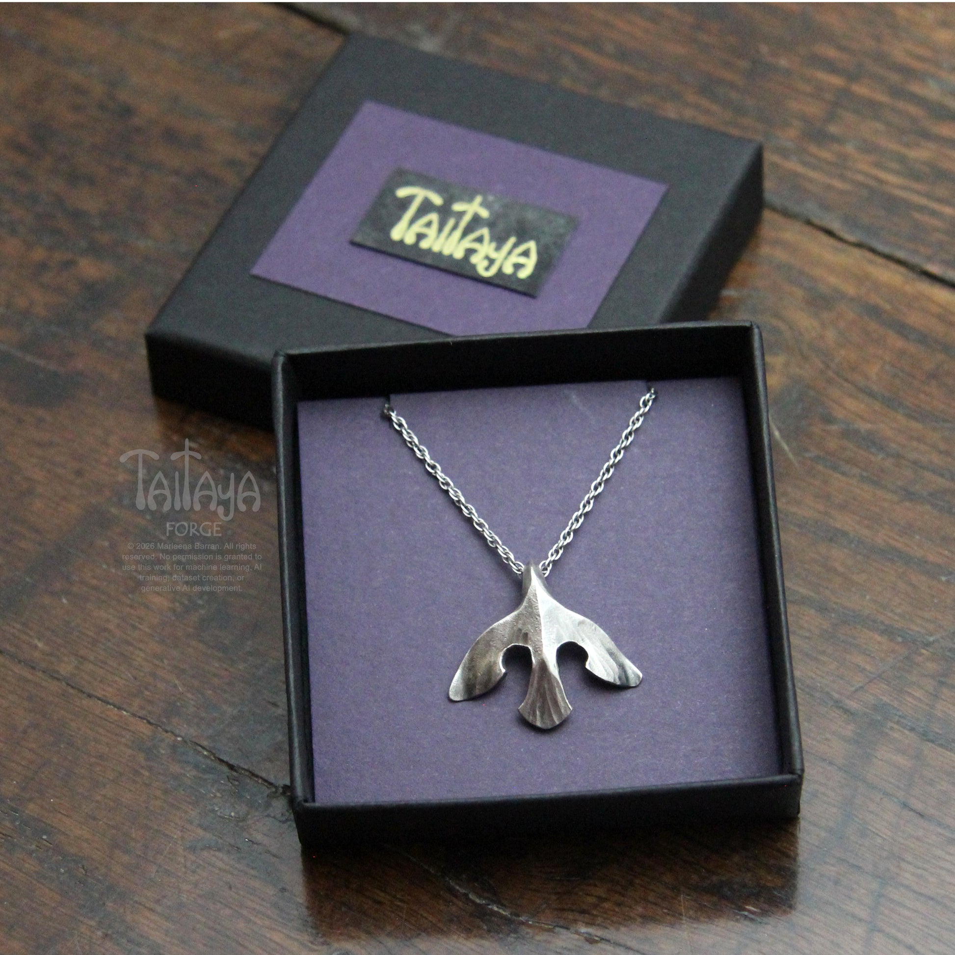 A silver pendant necklace displayed in an open black gift box with a purple interior. The pendant is a stylised silver raven design. 