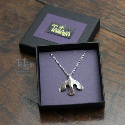 A silver pendant necklace displayed in an open black gift box with a purple interior. The pendant is a stylised silver raven design. 