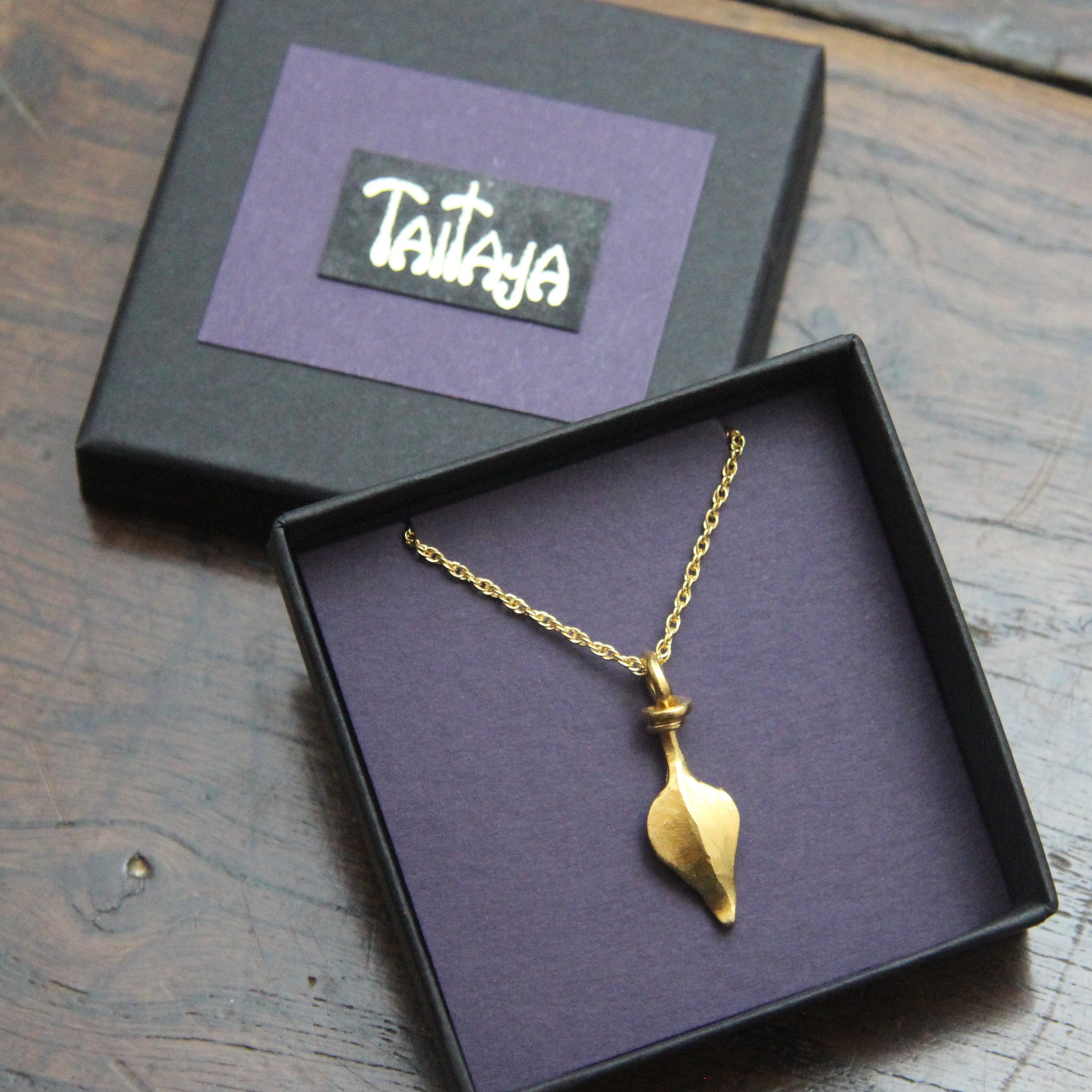 Gold necklace with a leaf pendant in a black and purple card jewelry box on a wooden surface, with 'Taitaya' branding.