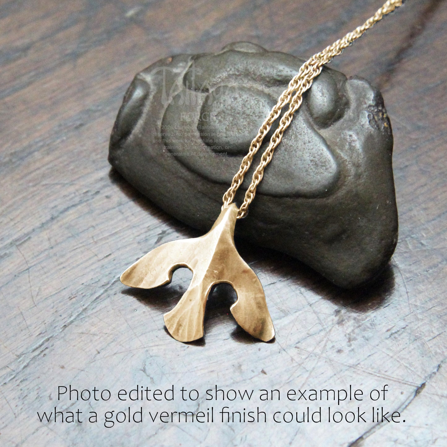 A mockup gold raven pendant necklace with a texture hangs from a gold chain, displayed on a dark gray stone over a wooden surface. Text at the bottom reads, 'Photo edited to show an example of what a gold vermeil finish could look like.