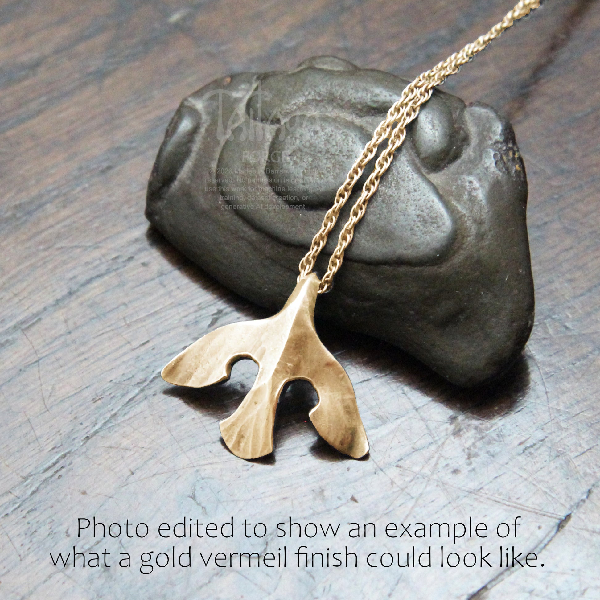 A mockup gold raven pendant necklace with a texture hangs from a gold chain, displayed on a dark gray stone over a wooden surface. Text at the bottom reads, 'Photo edited to show an example of what a gold vermeil finish could look like.