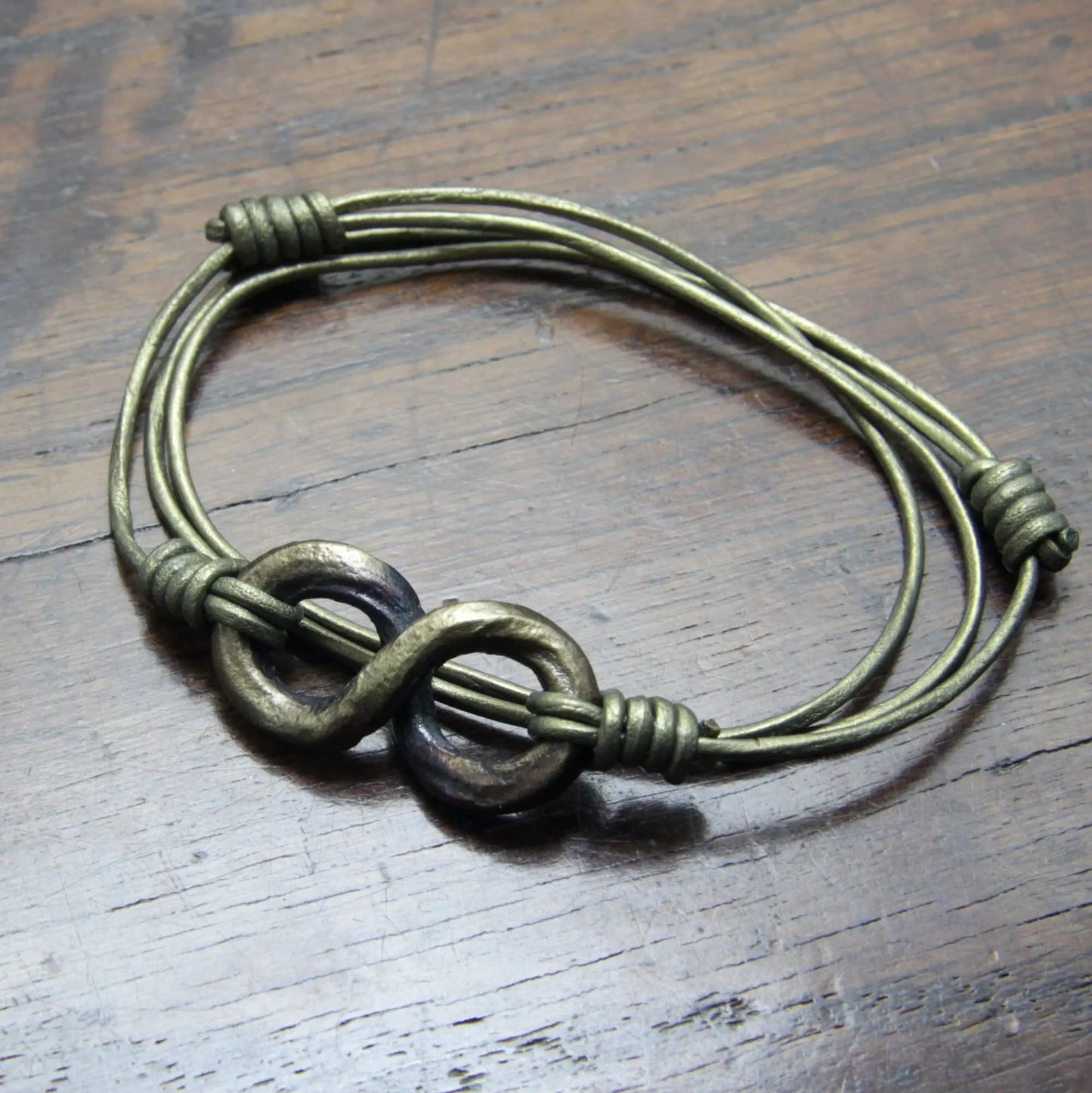 Main photo of a brass coloured Forged Iron Infinity Leather Wrap Bracelet and Necklace on a metallic green leather cord