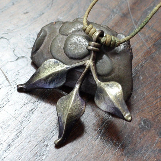 A close-up of a handmade triple leaf pendant necklace featuring a textured, dark metallic leaf design on a cord, resting on a scratched dark wooden surface.