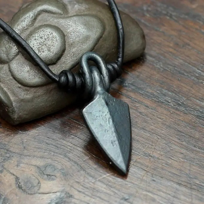 A dark metal arrowhead pendant necklace on a black cord, resting on a textured stone against a rustic wooden background.