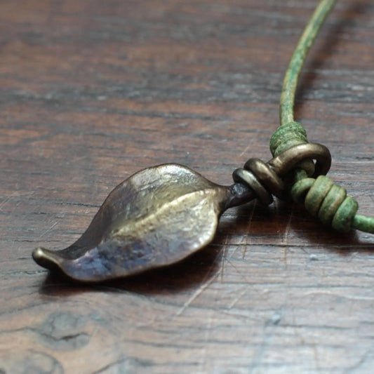 A close-up of a leaf-shaped pendant in an antique bronze finish, attached to a green knotted cord, resting on a dark wood surface.