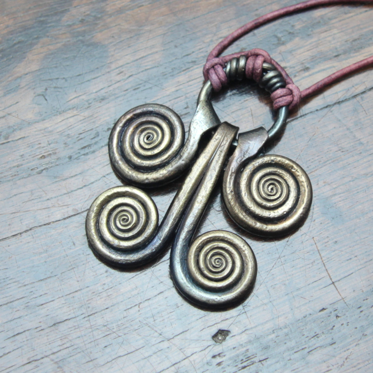 Aspiral pendant necklace with four coiled spirals and a central bar, wrapped in a dusty rose cord, shown on a weathered gray wooden surface.