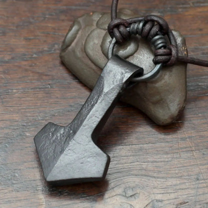 A dark gray forged iron Thors hammer pendant with a rustic, textured finish, attached to a dark brown leather cord and resting on a textured gray stone against a wooden background.
